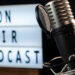 Großes Studiomikrofon mit Halterung vor einem Leuchtschild mit der Aufschrift „ON AIR PODCAST“.
