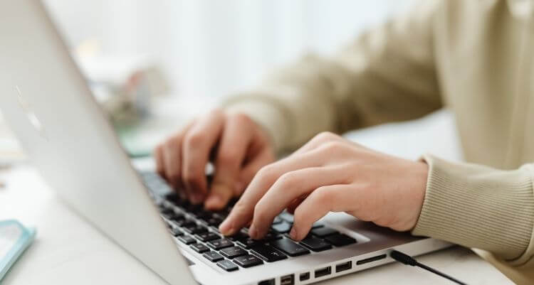 Hände tippen auf der Tastatur eines silberfarbenen Laptops, der auf einem Schreibtisch steht.