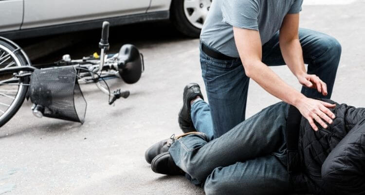 Person leistet Erste Hilfe für eine auf der Fahrbahn liegende Person; daneben liegt ein umgestürztes Fahrrad neben einem geparkten Fahrzeug.
