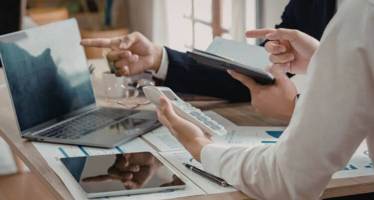 Arbeitsplatzszene mit mehreren Händen, die auf digitale Geräte, Dokumente und Diagramme zeigen.