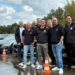 Gruppe von acht Personen in einheitlicher Unternehmenskleidung steht auf einem nassen Fahrtrainingsgelände vor mehreren geparkten Fahrzeugen und Verkehrsleitkegeln.