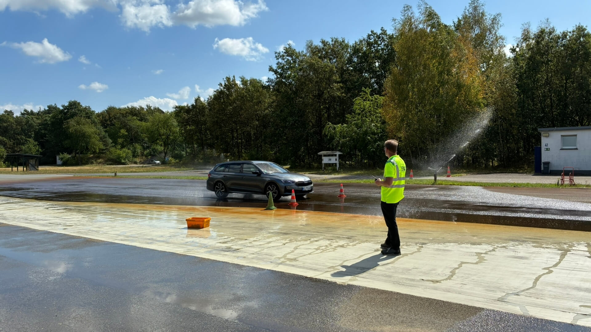 Pkw fährt über eine bewässerte Fahrbahn eines Fahrsicherheitszentrums, während eine Person in Warnkleidung den Ablauf der Übung beobachtet.