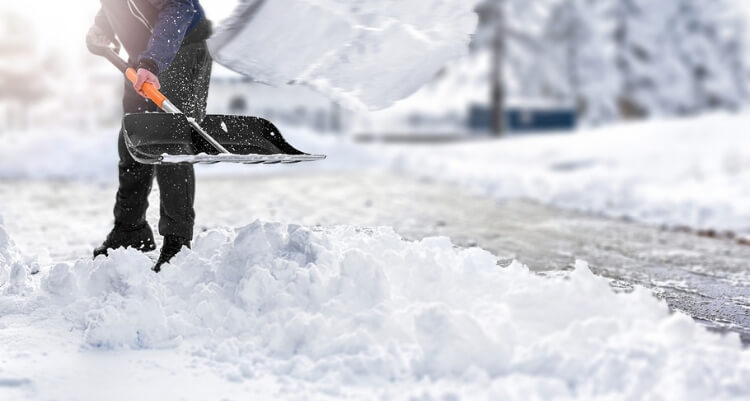 Person räumt mit einer Schneeschaufel eine verschneite Fläche und bewegt eine größere Schneemenge zur Seite.