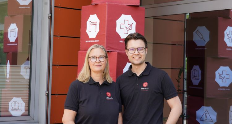 Zwei Personen in schwarzen Firmenpoloshirts mit Prevenio-Logo stehen vor roten Säulen mit Symbolen für Arbeitssicherheit und medizinische Eignung.