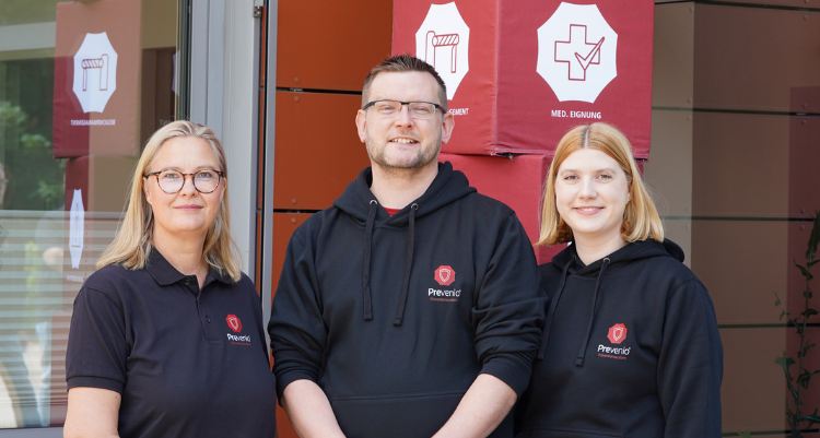 Drei Personen in schwarzen Prevenio-Hoodies und Poloshirts stehen vor roten Informationssäulen mit Symbolen zu Arbeitsschutz und Eignung.