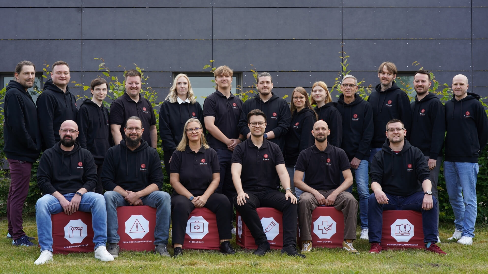 Großes Team in einheitlicher Arbeitskleidung, angeordnet in zwei Reihen vor einer modernen Gebäudefassade im Außenbereich.