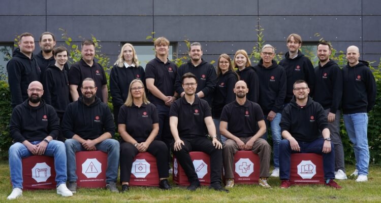 Gruppe von Mitarbeitern in einheitlichen dunklen Poloshirts, angeordnet in zwei Reihen vor einer Gebäudewand im Außenbereich.